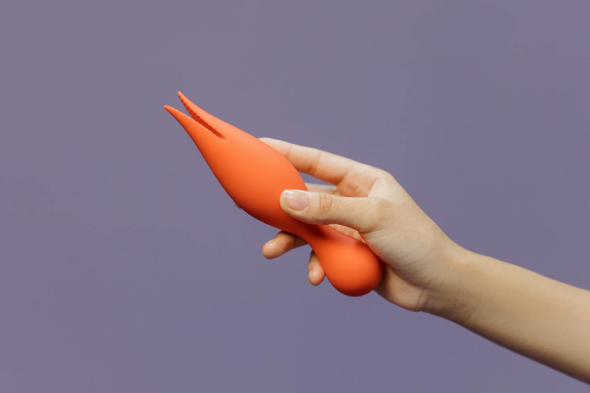 A close-up of a hand holding an orange vibrator against a minimalistic purple backdrop.